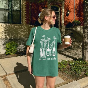 Peut inclure: Une femme portant un t-shirt vert avec un motif blanc de bouteilles de vin et de fleurs. Le t-shirt dit "la vie est belle".