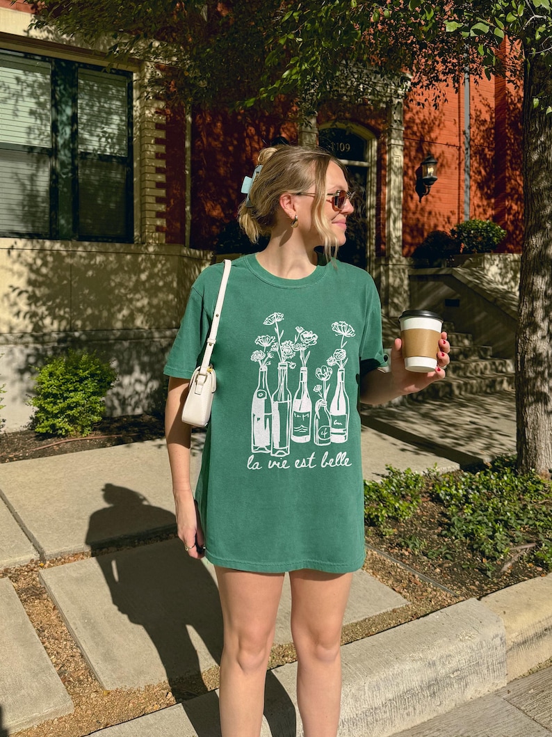 Peut inclure: Une femme portant un t-shirt vert avec un motif blanc de bouteilles de vin et de fleurs. Le t-shirt dit "la vie est belle".