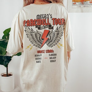 May include: A cream-colored t-shirt with a graphic print of a lightning bolt with wings. The graphic says "Mecan's Farewell Tour May 2024" and "Guest Stars: Ashley, Nicole, Kate, Claudia, Kelsey, Emily".