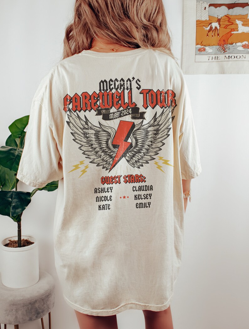 May include: A cream-colored t-shirt with a graphic print of a lightning bolt with wings. The graphic says "Mecan's Farewell Tour May 2024" and "Guest Stars: Ashley, Nicole, Kate, Claudia, Kelsey, Emily".