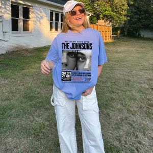 May include: A light blue t-shirt with a graphic print of a wedding announcement for "The Johnsons" with the text "Wedding Tour 2025" and "David & Anna". The announcement also includes the date "24 May" and the location "Park City".