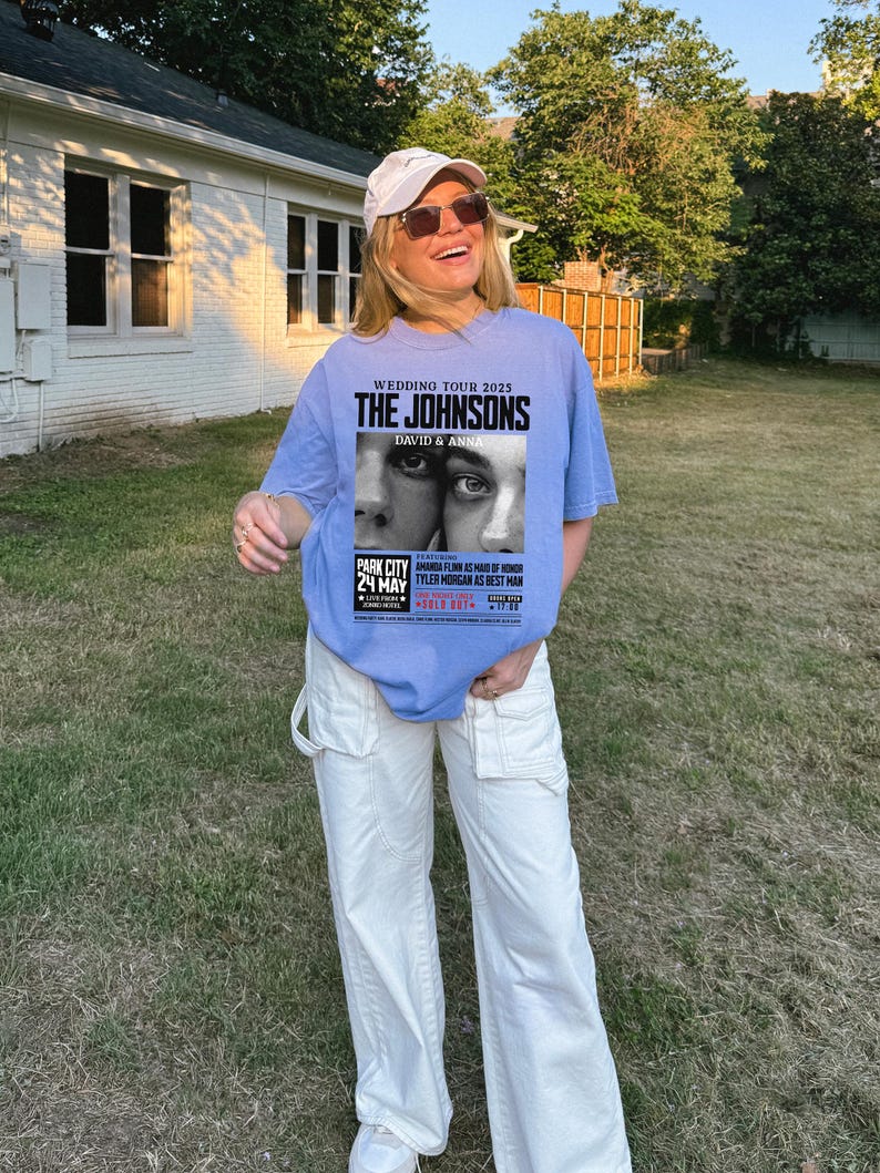 May include: A light blue t-shirt with a graphic print of a wedding announcement for "The Johnsons" with the text "Wedding Tour 2025" and "David & Anna". The announcement also includes the date "24 May" and the location "Park City".