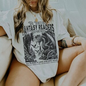 May include: A white t-shirt with a black graphic print of a dragon and the text "The Torture Fantasy Readers Department" and "All's Fair in Love and Epic Tales".