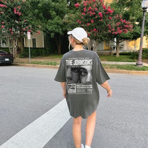 May include: A gray oversized t-shirt with a black and white graphic print of a wedding announcement for "The Johnsons" with the text "Wedding Tour 2025" and "David & Anha". The graphic also includes a black and white photo of a couple and the text "Park City 24 May".