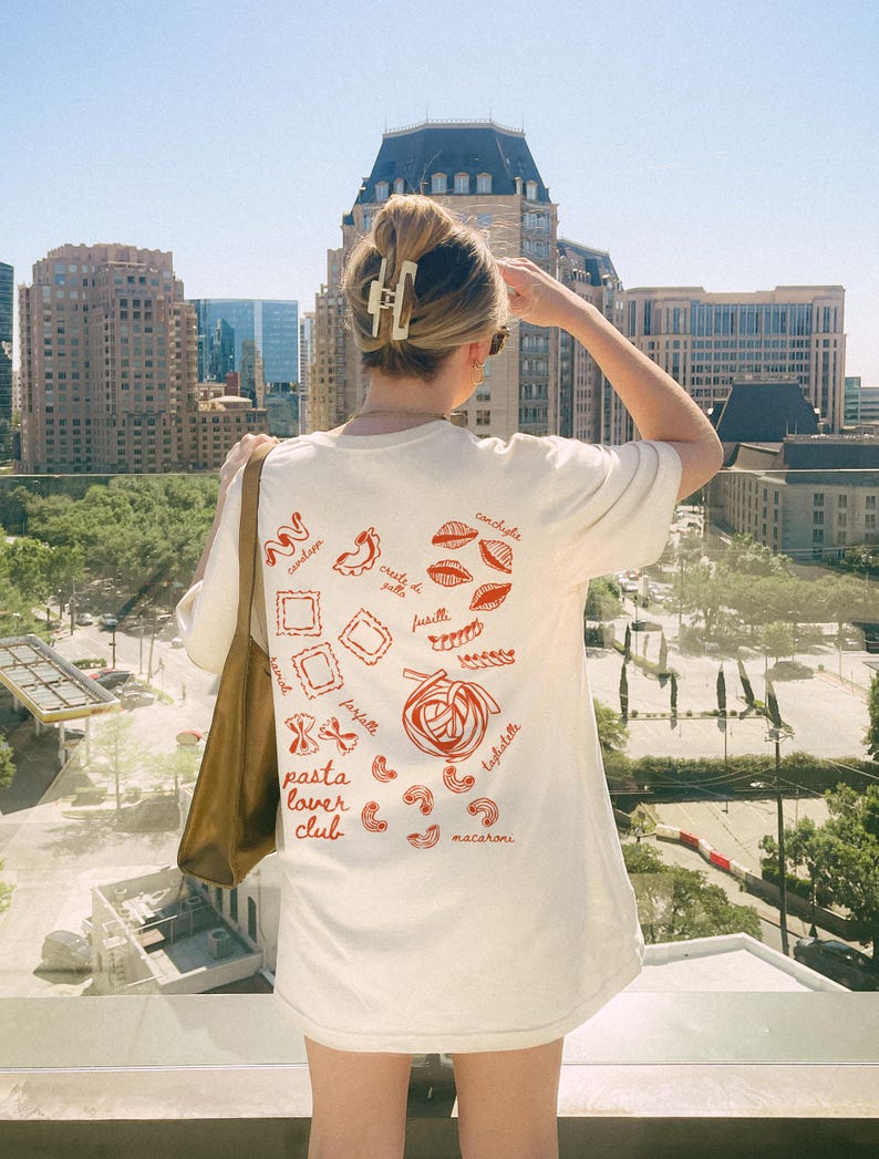 Puede incluir: Camiseta blanca con un gr&aacute;fico rojo de diferentes tipos de pasta, incluyendo espaguetis, macarrones y fusilli. El texto "pasta lover club" est&aacute; impreso en la camiseta.