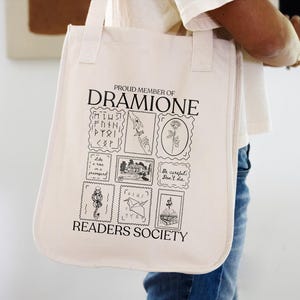 May include: A white canvas tote bag with black text that reads "Proud Member of Dramione Readers Society". The bag features a series of black and white illustrations of stamps with various designs, including a rose, a house, a bird, and a snake.
