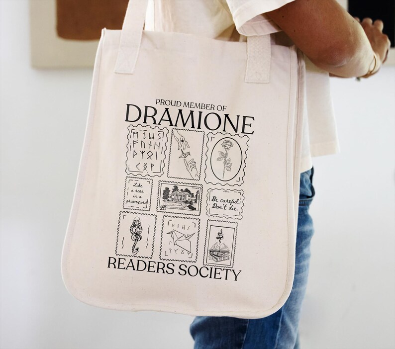 May include: A white canvas tote bag with black text that reads "Proud Member of Dramione Readers Society". The bag features a series of black and white illustrations of stamps with various designs, including a rose, a house, a bird, and a snake.