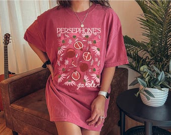 May include: A maroon-colored t-shirt dress with the text "PERSEPHONE'S garden" and a floral and pomegranate design. The design features pink flowers and sliced pomegranates. The dress is a loose fit.