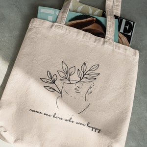 May include: A beige tote bag with a black line drawing of a bust with leaves growing out of the top. The text "name one hero who was happy" is printed below the drawing.