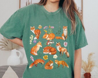 May include: A green t-shirt with a print of multiple orange foxes and flowers. The foxes are in various poses, including sleeping, sitting, and standing.