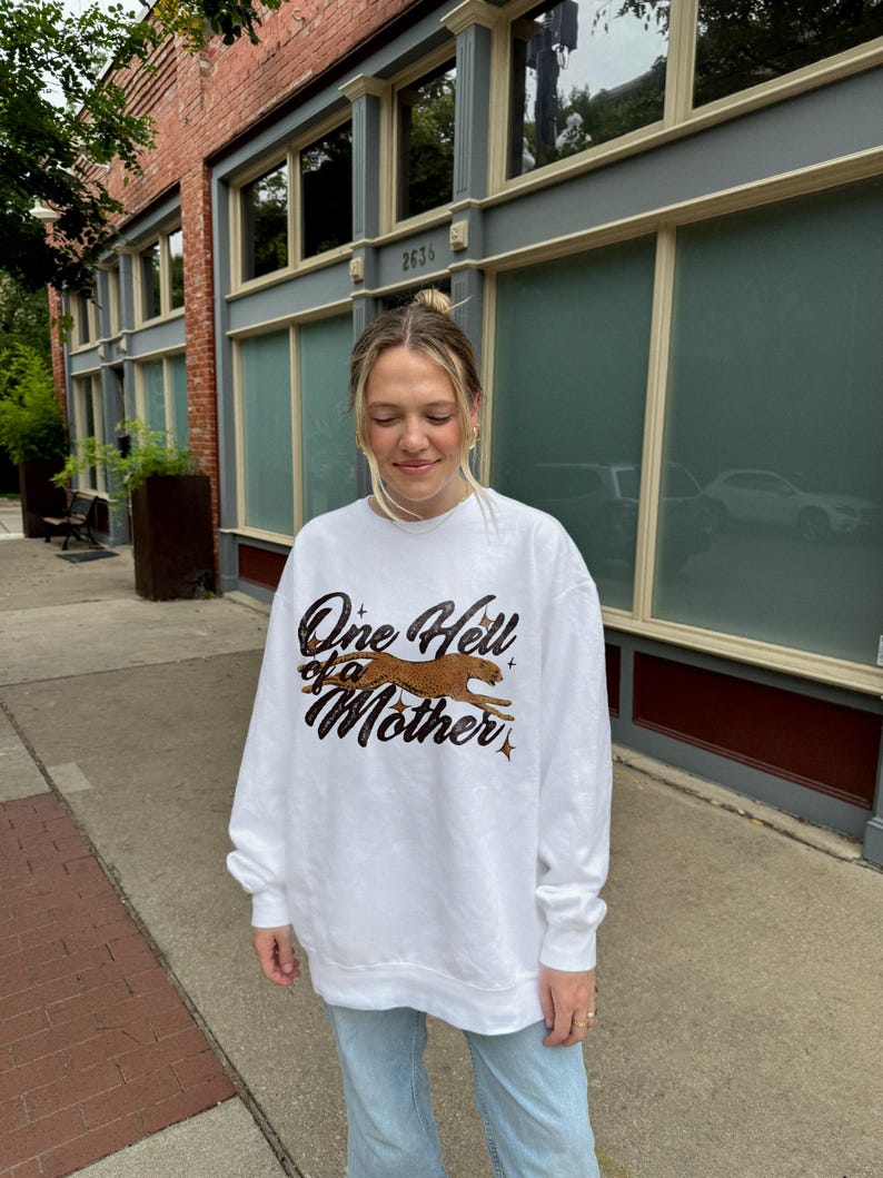 Peut inclure: Sweat-shirt blanc avec un imprim&eacute; graphique d'un gu&eacute;pard courant et le texte "One Hell of a Mother".