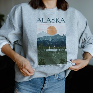 Pu&ograve; includere: Felpa grigia con una stampa grafica di un paesaggio montano con un sole e il testo "ALASKA LAND OF THE MIDNIGHT SUN".