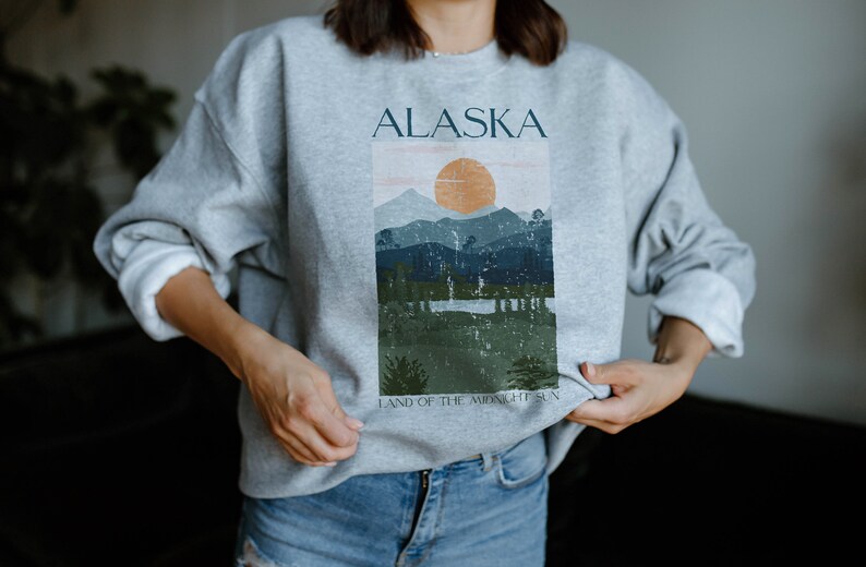 Pu&ograve; includere: Felpa grigia con una stampa grafica di un paesaggio montano con un sole e il testo "ALASKA LAND OF THE MIDNIGHT SUN".