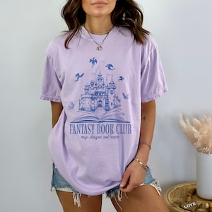 May include: A lavender t-shirt with a blue graphic of a castle, book, dragons, and butterflies. The text "Fantasy Book Club" and "magic, dragons and fairies" is printed on the shirt.
