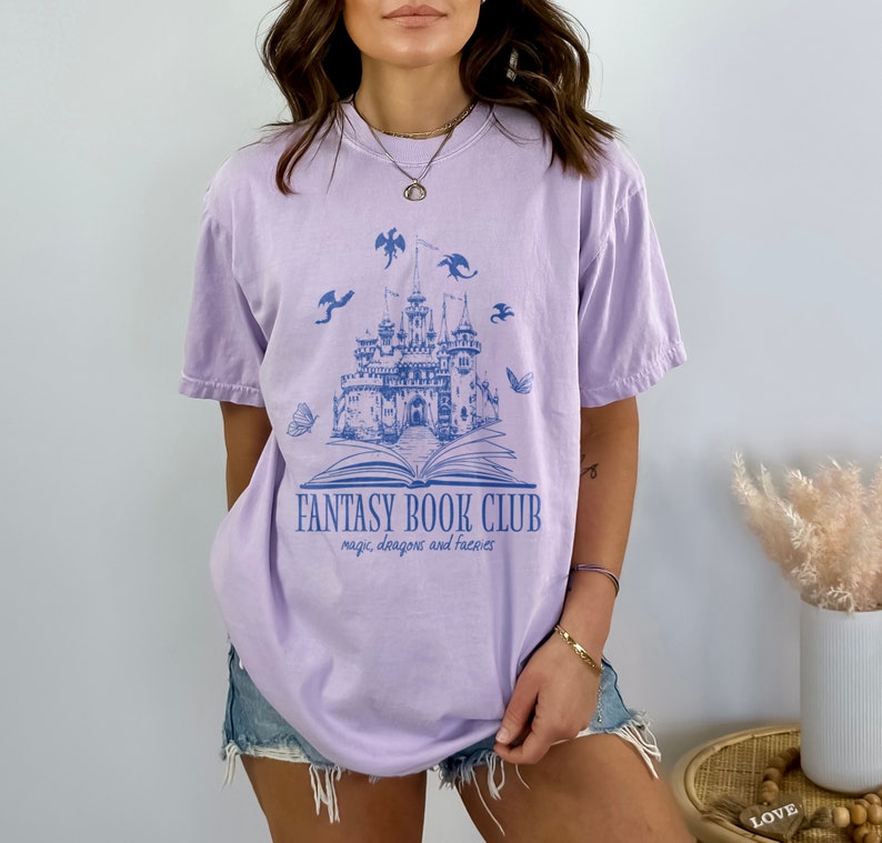 May include: A lavender t-shirt with a blue graphic of a castle, book, dragons, and butterflies. The text "Fantasy Book Club" and "magic, dragons and fairies" is printed on the shirt.