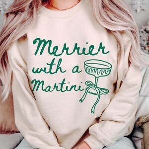 May include: A cream colored sweatshirt with the text "Merrier with a Martini" in green lettering. A green martini glass with a green bow is pictured next to the text.
