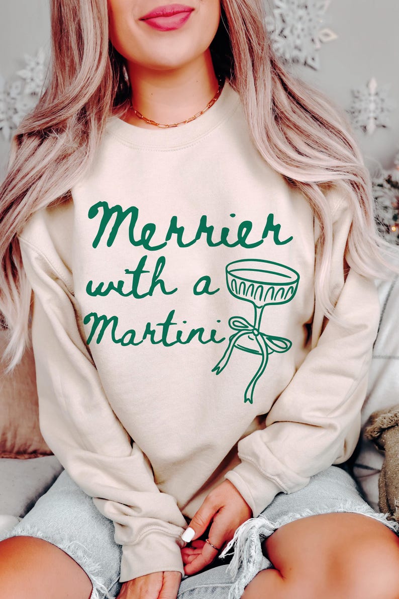 May include: A cream colored sweatshirt with the text "Merrier with a Martini" in green lettering. A green martini glass with a green bow is pictured next to the text.