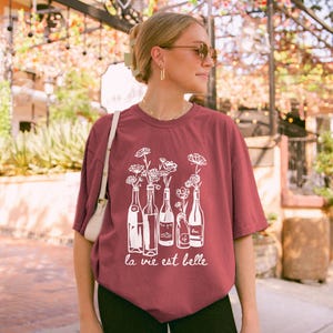 Peut inclure: Un t-shirt marron avec un motif blanc de bouteilles de vin avec des fleurs &agrave; l'int&eacute;rieur. Le texte "la vie est belle" est imprim&eacute; sous le motif.