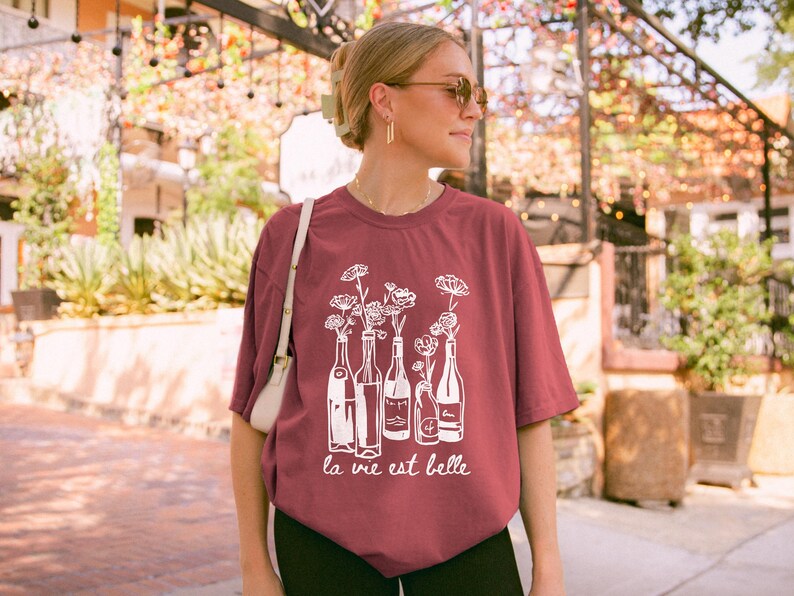 Peut inclure: Un t-shirt marron avec un motif blanc de bouteilles de vin avec des fleurs &agrave; l'int&eacute;rieur. Le texte "la vie est belle" est imprim&eacute; sous le motif.