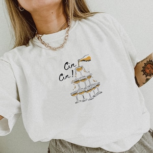 Könnte beinhalten: Weißes T-Shirt mit einem Aufdruck eines Champagnerturms und dem Text "Cin Cin!"