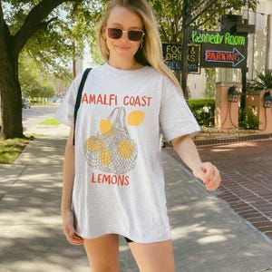 May include: A grey t-shirt with a graphic of lemons in a mesh bag. The text "Amalfi Coast" and "Lemons" is printed on the shirt.