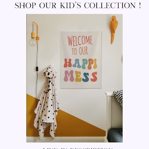 May include: A white canvas wall hanging with the text "Welcome to our Happy Mess" in colourful, bubble-like letters. The wall is painted yellow and white, and there is a black and white polka dot blanket hanging on a hook.