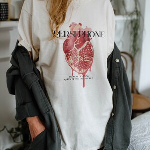 May include: White t-shirt with a graphic of a heart-shaped pomegranate and the text "PERSEPHONE, GODDESS OF SPRING, QUEEN OF THE UNDERWORLD".