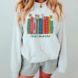 Op de afbeelding: Een grijs sweatshirt met een afbeelding van een stapel boeken met bloemmotieven en de tekst "Nicole's Book Club".