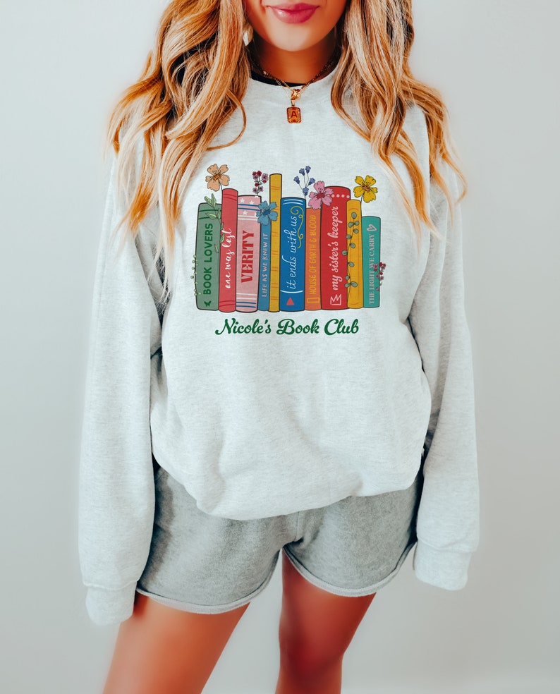 Op de afbeelding: Een grijs sweatshirt met een afbeelding van een stapel boeken met bloemmotieven en de tekst "Nicole's Book Club".