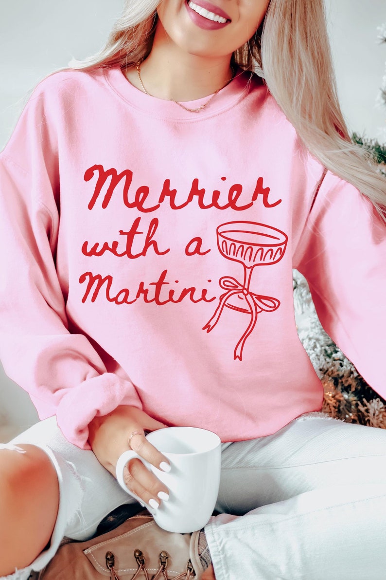 May include: Pink sweatshirt with the text "Merrier with a Martini" in red lettering and a red illustration of a martini glass with a bow.
