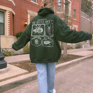 K&ouml;nnte beinhalten: Ein dunkelgr&uuml;ner Hoodie mit einem wei&szlig;en Aufdruck von K&auml;fern und dem Text "Proud Member of The Beetle Society".