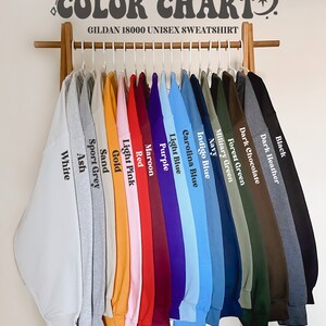 May include: A color chart of Gildan 18000 unisex sweatshirts. The chart shows 16 different colors, including white, ash, sport grey, sand, gold, light pink, red, maroon, purple, light blue, carolina blue, indigo blue, navy, military green, forest green, dark chocolate, dark heather, and black.
