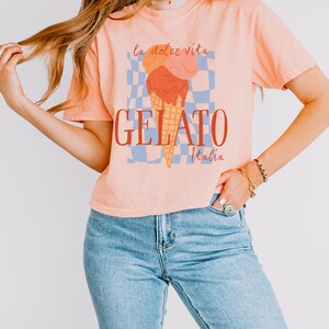 May include: Peach-colored t-shirt with a gelato graphic. The graphic features an ice cream cone with three scoops in orange, pink, and red, with the words "GELATO Italia" below. The phrase "la dolce vita" is above the ice cream.