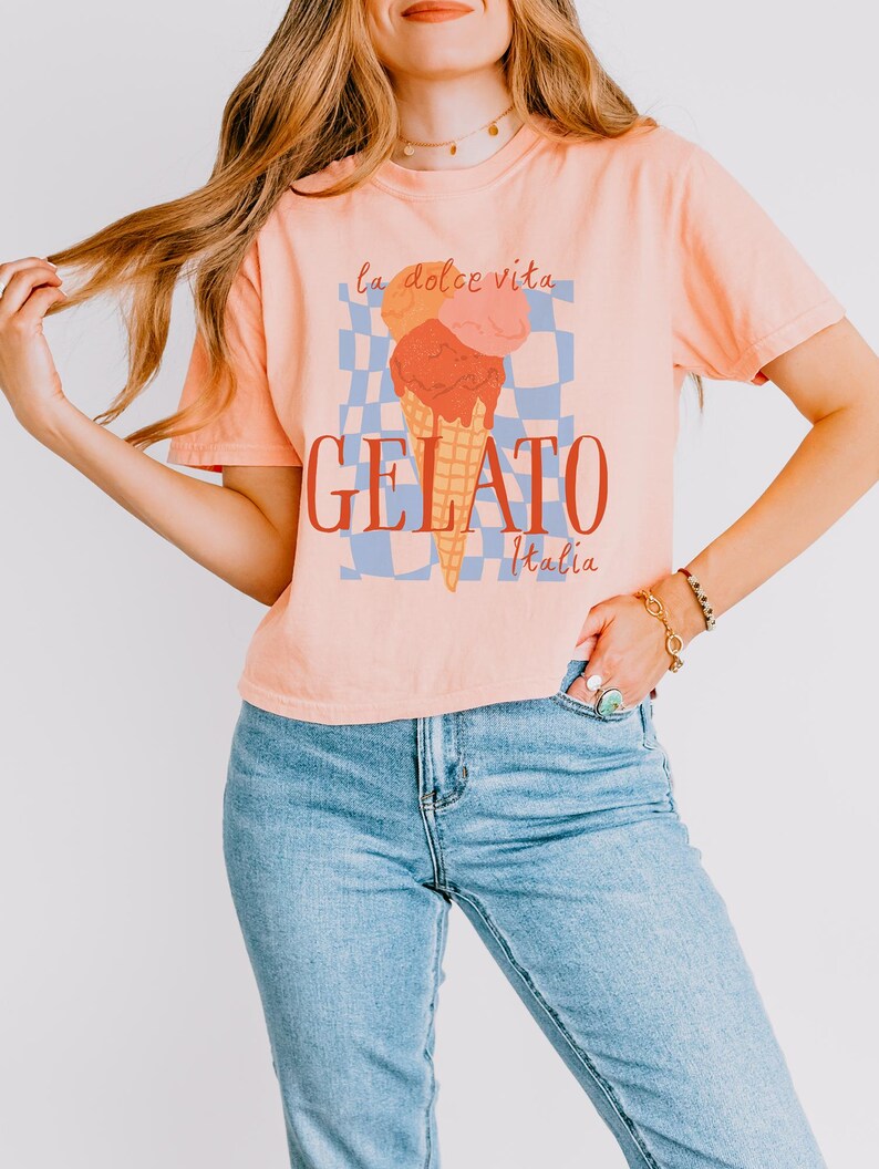 May include: Peach-colored t-shirt with a gelato graphic. The graphic features an ice cream cone with three scoops in orange, pink, and red, with the words "GELATO Italia" below. The phrase "la dolce vita" is above the ice cream.