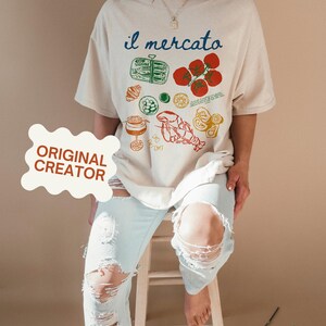 May include: A beige oversized t-shirt with a graphic print of various Italian food items, including tomatoes, lemons, pasta, and a lobster. The text "il mercato" is printed in blue on the shirt.