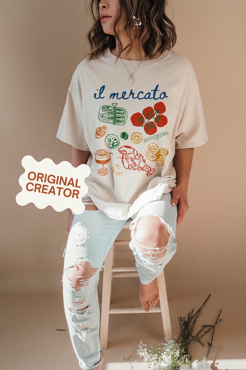 May include: A beige oversized t-shirt with a graphic print of various Italian food items, including tomatoes, lemons, pasta, and a lobster. The text "il mercato" is printed in blue on the shirt.