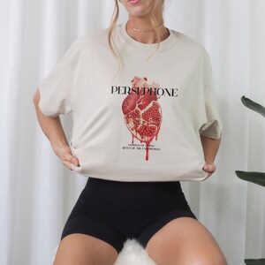 May include: A beige t-shirt with a graphic of a red heart with a pomegranate inside. The text "PERSEPHONE" is above the heart and "GODDESS OF SPRING QUEEN OF THE UNDERWORLD" is below the heart.
