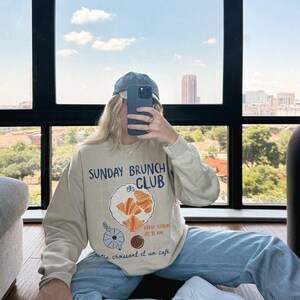 May include: A beige sweatshirt with a graphic print of a plate of croissants and the text "Sunday Brunch Club" and "Every Sunday at 11 am Un croissant et un cafe".