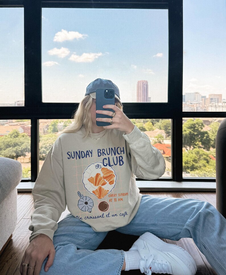 May include: A beige sweatshirt with a graphic print of a plate of croissants and the text "Sunday Brunch Club" and "Every Sunday at 11 am Un croissant et un cafe".