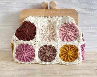 May include: A cream-colored crochet clutch with a wooden frame and clasp. The front features a patchwork design of colorful circular patterns in shades of brown, pink, and yellow. The bag is a stylish accessory.