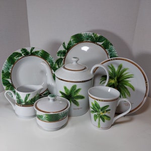 May include: A white teapot with a green and brown tropical leaf design, a white sugar bowl with a green and brown tropical leaf design, a white creamer with a green and brown tropical leaf design, and two white mugs with green and brown tropical leaf designs. The teapot has a handle and a spout. The sugar bowl has a lid. The creamer has a handle. The mugs have handles. All of the pieces are on a white surface.