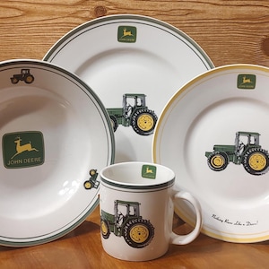 May include: A white ceramic dinnerware set featuring a green tractor design and the John Deere logo. The set includes plates and a mug. The plates have green or yellow rims. The mug has a handle.
