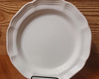 May include: White ceramic dinner plate with a scalloped edge. The plate is on a wooden surface.
