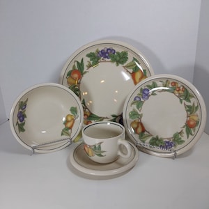 Op de afbeelding: Een set van vier crèmekleurige keramische schalen met een fruit- en wijnstokontwerp. De set bevat een groot dinerbord, een kleiner saladebord, een kom en een theekopje met schoteltje.