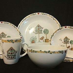 May include: A set of white ceramic dishes with a birdhouse and floral design. The set includes a large plate, a smaller plate, a bowl, and a mug.