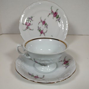 May include: A white teacup and saucer set with a floral design. The cup and saucer have a gold trim. The floral design features pink roses and green leaves.
