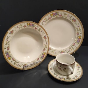 May include: A set of white porcelain dinnerware with a floral pattern in shades of pink, yellow, and green. The set includes a dinner plate, a soup bowl, a teacup, and a saucer. The edges of the plates and bowls are trimmed with a thin gold band.