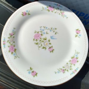May include: White ceramic plate with a gold rim and a floral design featuring pink and white flowers, green leaves, and a blue bird.