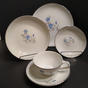 Può includere: Un set di stoviglie in porcellana bianca con un motivo di rose blu e un bordo argentato. Il set comprende un piatto da portata, un piatto da insalata, una ciotola per la zuppa e una tazza da tè con piattino.