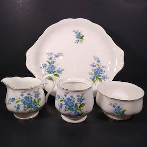 Może przedstawiać: Biały porcelanowy zestaw do herbaty z kwiatowym wzorem niezapominajek. Zestaw zawiera dzbanek do śmietanki, cukiernicę i tacę do serwowania.